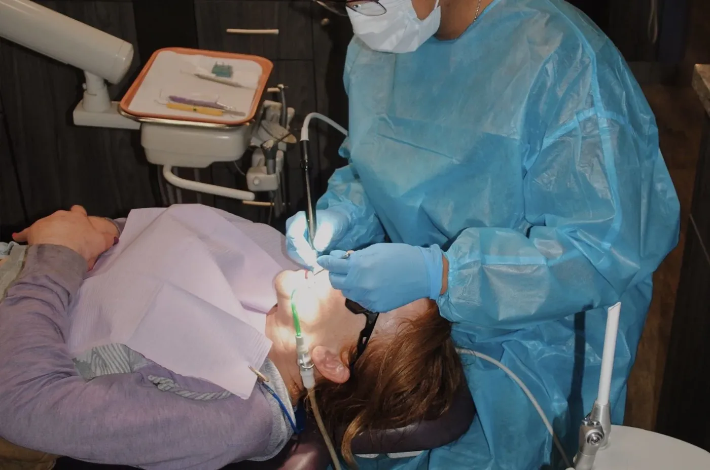 Dentist in a blue protective gown and mask treating a patient in the dental chair
