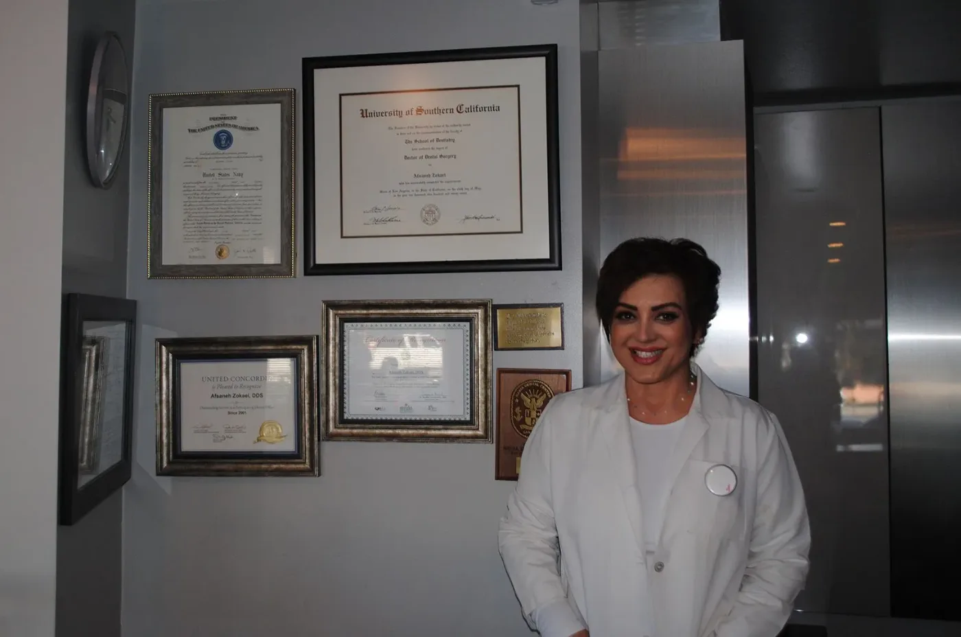 Dr. Zokaei in a white coat standing beside her framed diplomas