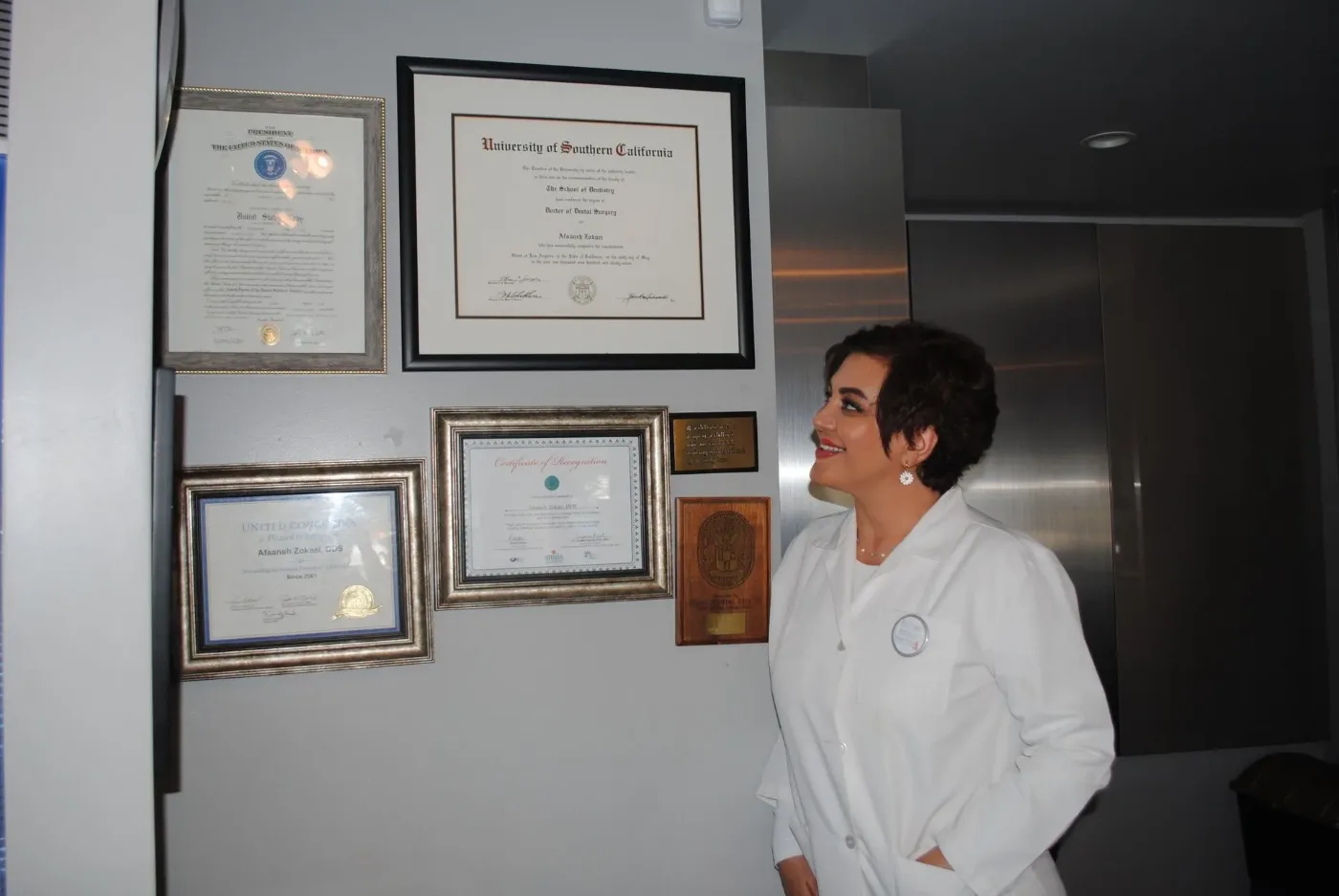 Dr. Zokaei in a white coat looking at her framed diplomas and certificates